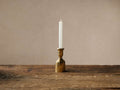 Textured brass Thira Taper Holder with a lit white taper candle, on a rustic wooden table against a neutral wall.