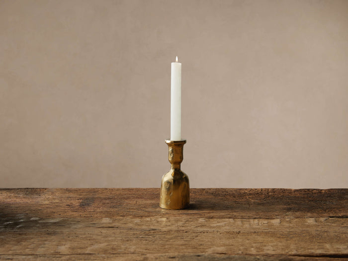 Textured brass Thira Taper Holder with a lit white taper candle, on a rustic wooden table against a neutral wall.
