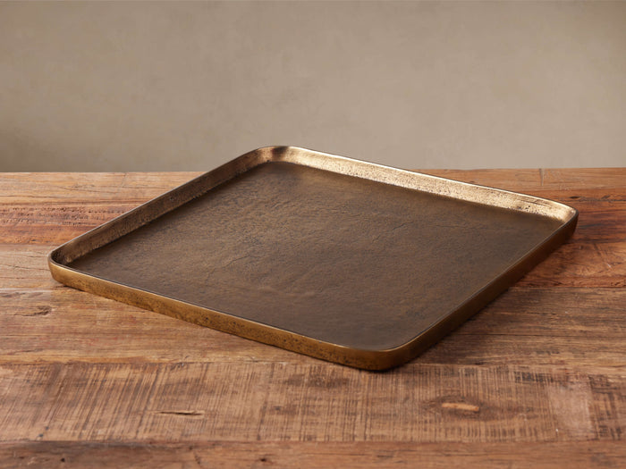 Tova Tray, square antique brass-finished cast aluminum tray with rustic texture, angled on a wooden surface.
