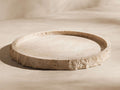 Round, light beige travertine bath tray with rough, natural stone edges and textured surface, angled on a creamy background.