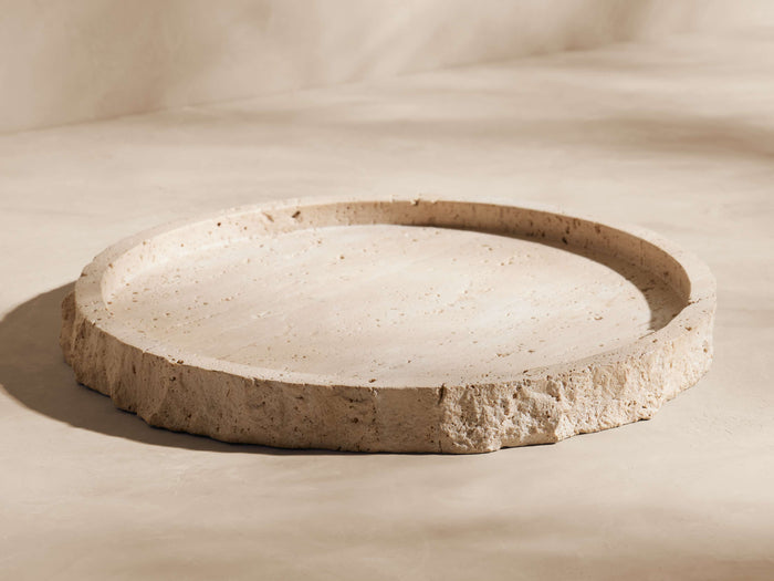Round, light beige travertine bath tray with rough, natural stone edges and textured surface, angled on a creamy background.