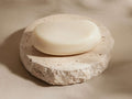 Round, textured beige travertine soap dish with a creamy white oval soap bar resting on top, set against a soft beige background.