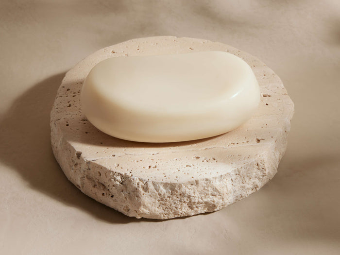 Round, textured beige travertine soap dish with a creamy white oval soap bar resting on top, set against a soft beige background.
