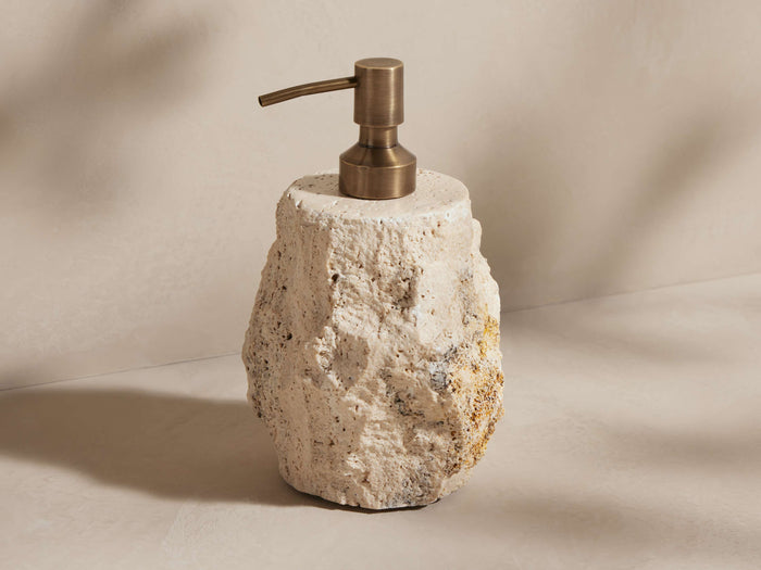 Cream travertine soap pump with rough texture and natural veining, topped with an antique brass pump, standing on a light surface.