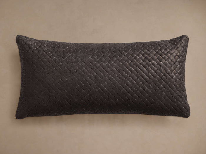 Charcoal grey Treccia Woven Lumbar Pillow, horizontal, featuring an intricate basket-weave leather texture on a tan wall.