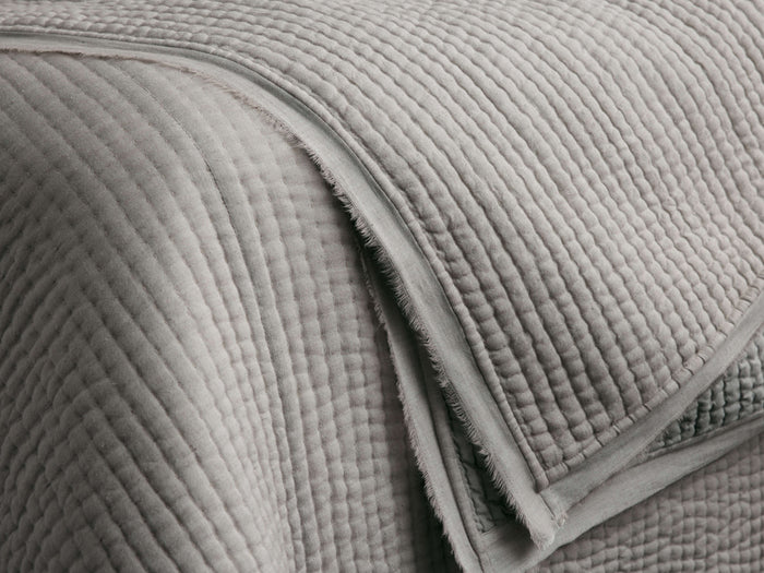 Close-up of a light gray Tulaya Cotton Quilt, folded with parallel stitching, showing the soft texture and frayed edge detail.