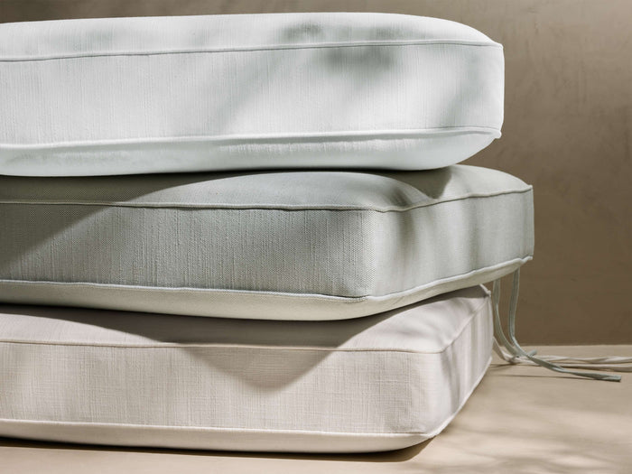 Stacked white, sage green, and beige Van Outdoor Water-Repellent Replacement Cushions with piping, sunlit on textured wall background.