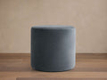 Velvet Round Pouf in a rich gray color on a wooden floor. The pouf is cylindrical with a smooth, velvety texture and stands against a light beige wall.
