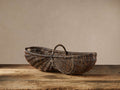 Handwoven vintage-stained arurog reed basket with a handle, resting on a rustic wooden surface against a light wall.