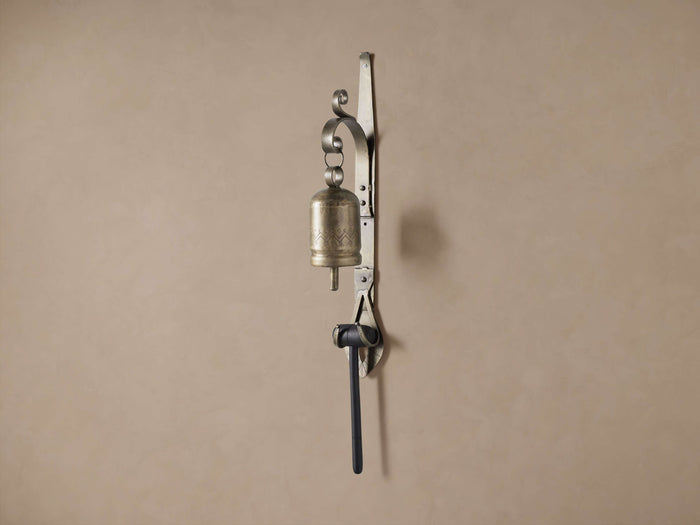 Wall-mounted antique brass dinner bell with etched patterns, dark mallet, and scrolled iron bracket against a plain wall.