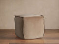 Square, light tan washed velvet pouf with prominent flanged edges, sitting on a warm wooden floor against a light beige wall.