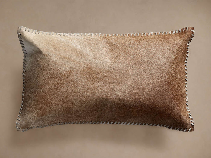 Tan hide lumbar pillow with varied brown and tan fur texture, rectangular shape, and white whipstitching along its edges.