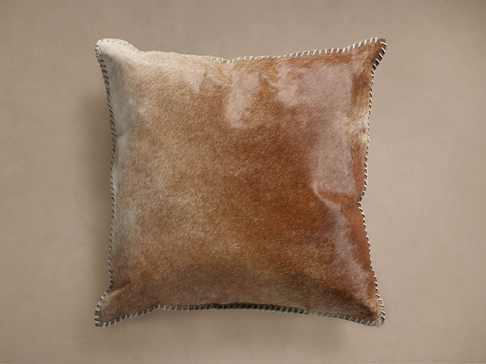 Square brown hide pillow with varied shades, featuring white whipstitch edges, shown slightly angled on a light brown background.