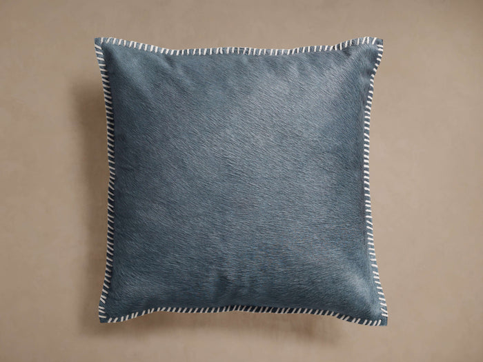 Front view of a storm blue square throw pillow with visible cowhide texture and white whipstitch trim around the edges.