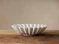 White marble Ruffle Bowl with wavy edges, handcrafted, sitting on a rustic wooden surface against a light beige wall.