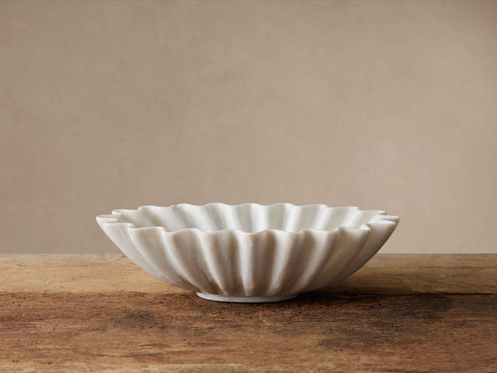 White marble Ruffle Bowl with wavy edges, handcrafted, sitting on a rustic wooden surface against a light beige wall.