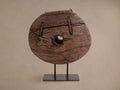 Rustic teak cart wheel with a central hole, showing visible grain and cracks, standing upright on a dark iron display stand.