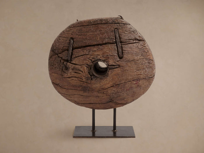 Rustic teak cart wheel with a central hole, showing visible grain and cracks, standing upright on a dark iron display stand.