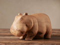 Hand-carved light brown Wooden Hippo figurine standing on a rustic wood surface, facing slightly left. Natural wood grain visible.