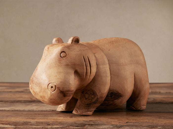 Hand-carved light brown Wooden Hippo figurine standing on a rustic wood surface, facing slightly left. Natural wood grain visible.