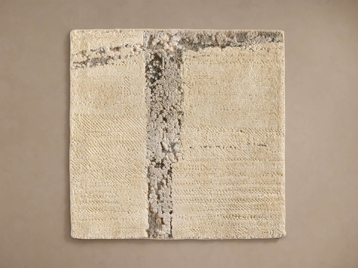 A square beige rug with a vertical textured stripe in darker shades and intricate patterns, laid on a neutral background.