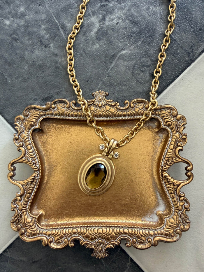 An ornate antique-style gold pendant necklace featuring a decorative tray-shaped frame with a central oval amber gemstone, displayed on a dark gray stone surface.
