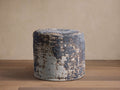 Cylindrical Zaikin Pouf featuring a distressed jacquard pattern in dark blue, light blue, and beige on a wood floor.