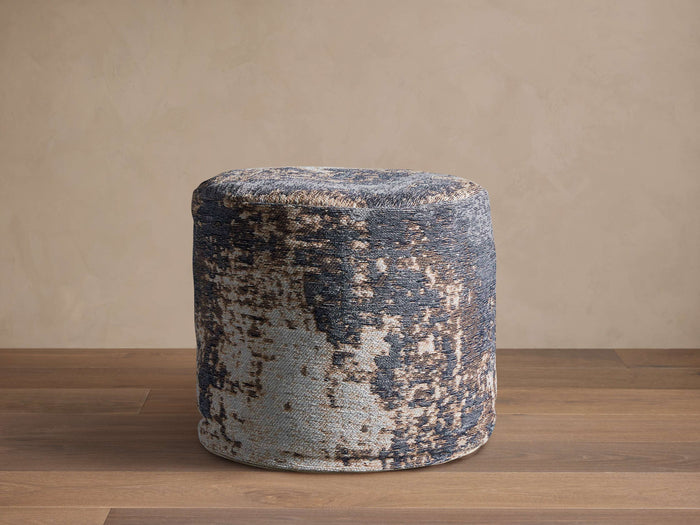 Cylindrical Zaikin Pouf featuring a distressed jacquard pattern in dark blue, light blue, and beige on a wood floor.