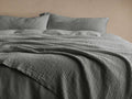 Grey Zehra Linen Bed Blanket draped softly over a bed with matching pillows; features a textured, gauzy surface with stitched details.
