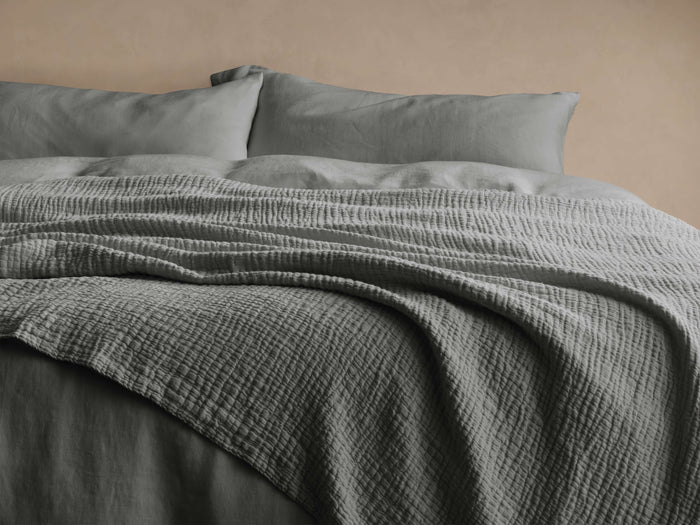 Grey Zehra Linen Bed Blanket draped softly over a bed with matching pillows; features a textured, gauzy surface with stitched details.