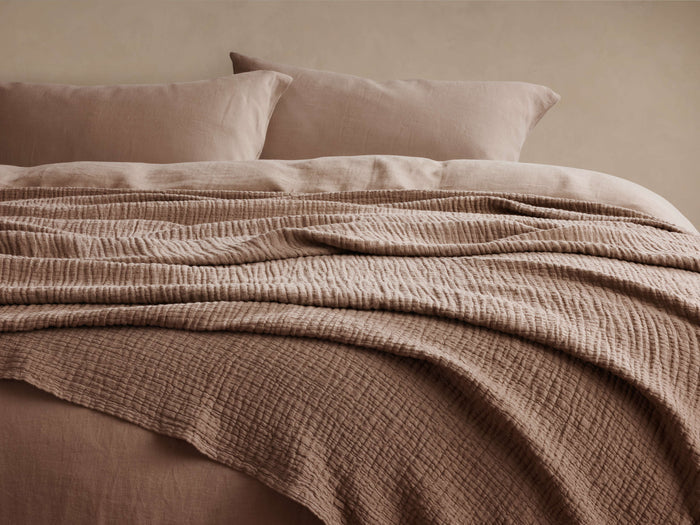 Zehra Clay Linen Bed Blanket draped over a bed, revealing its soft, gauzy, crinkled texture. Clay-colored pillows behind.