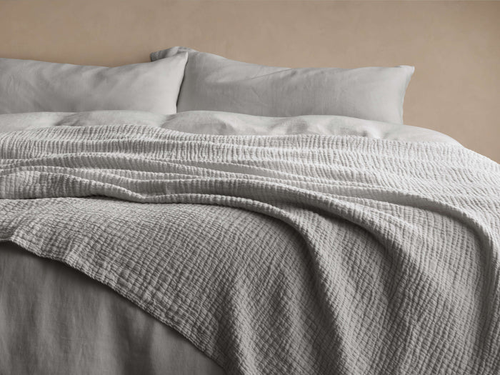 Light grey Zehra Linen Bed Blanket with a textured, gauzy weave, draped over a bed with matching pillows against a beige wall.
