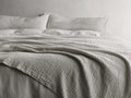 Zehra Linen Bed Blanket draped over a bed, showing its soft, gauzy texture and double needle stitching in a neutral tone.