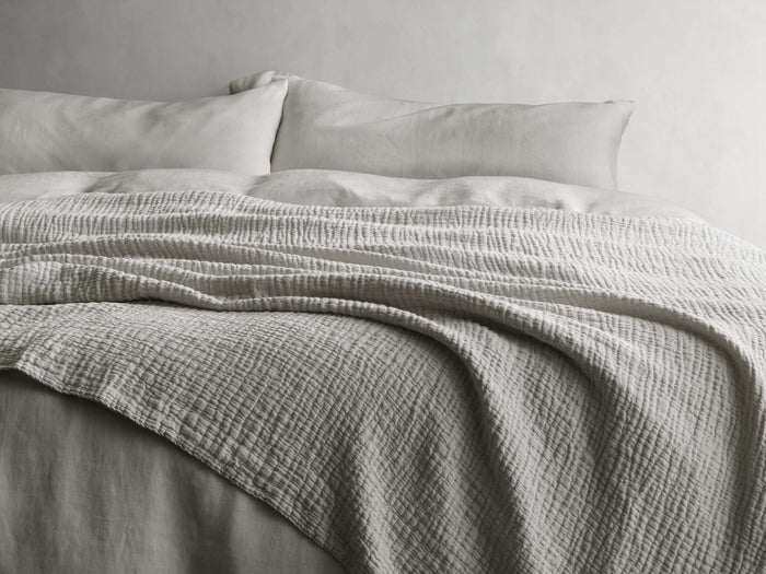 Zehra Linen Bed Blanket draped over a bed, showing its soft, gauzy texture and double needle stitching in a neutral tone.