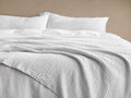 White Zehra Linen Bed Blanket draped softly over a bed, showcasing its crinkled, gauzy texture. White pillows in background.