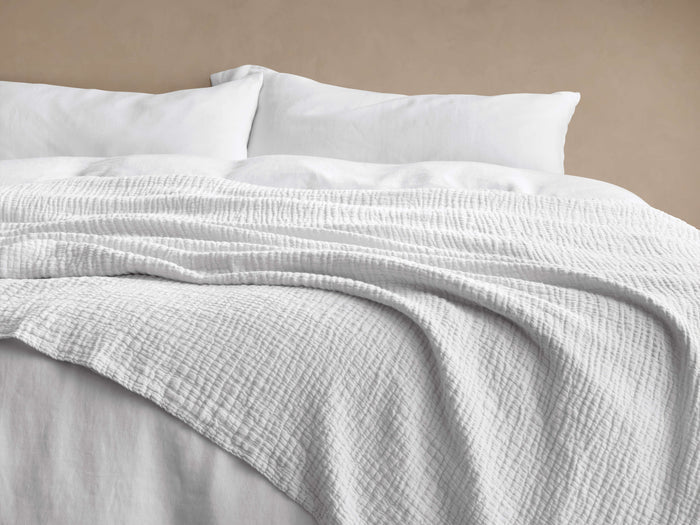 White Zehra Linen Bed Blanket draped softly over a bed, showcasing its crinkled, gauzy texture. White pillows in background.