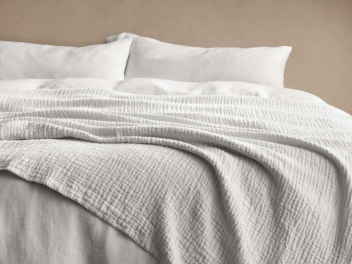 Light off-white Zehra Linen Bed Blanket draped softly on a bed with white pillows, showing its crinkled, gauzy texture.
