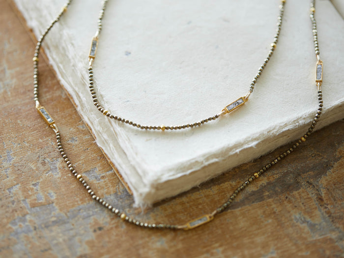 Ziya Necklace with small round pyrite beads and rectangular gold-plated settings; placed on a textured surface.