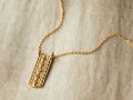Zuma Necklace with a yellow bronze rectangular pendant, centered white diamond, on a 14kt gold-fill chain, displayed on fabric.